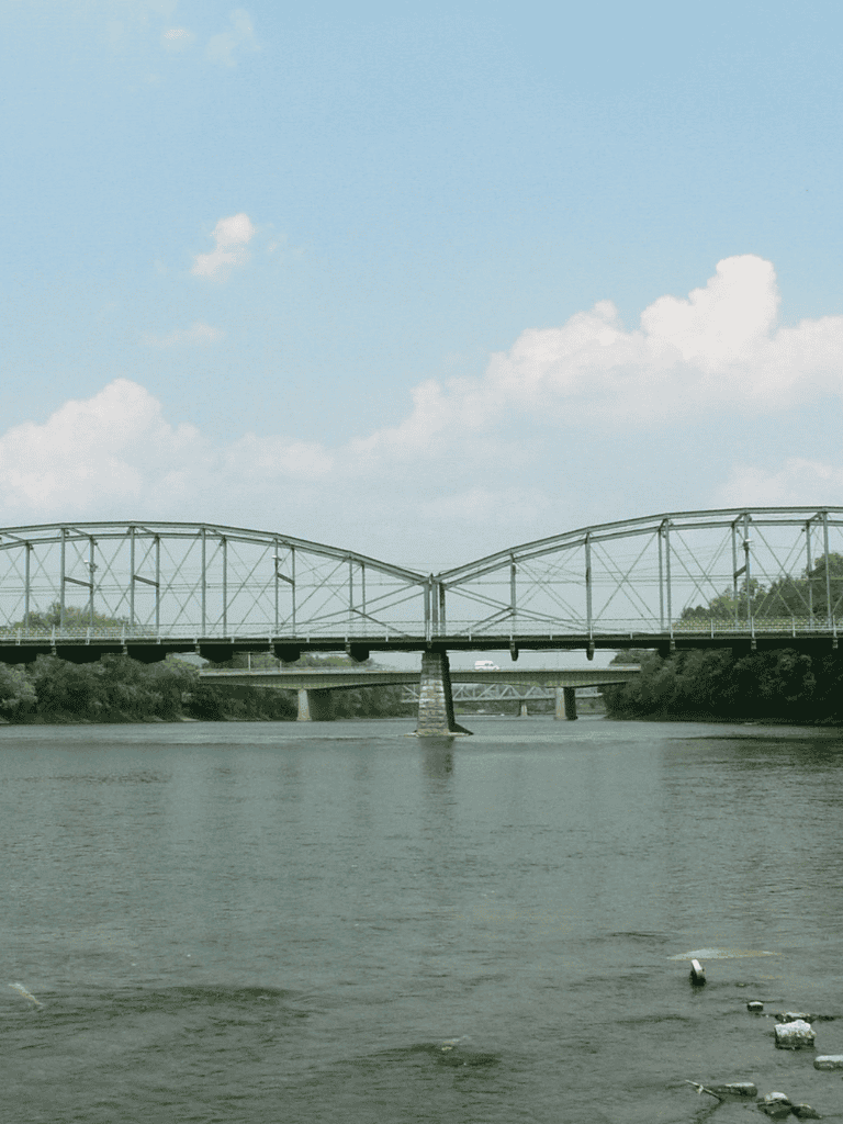 Restored steel bridge over river with lush green trees and blue sky in background.