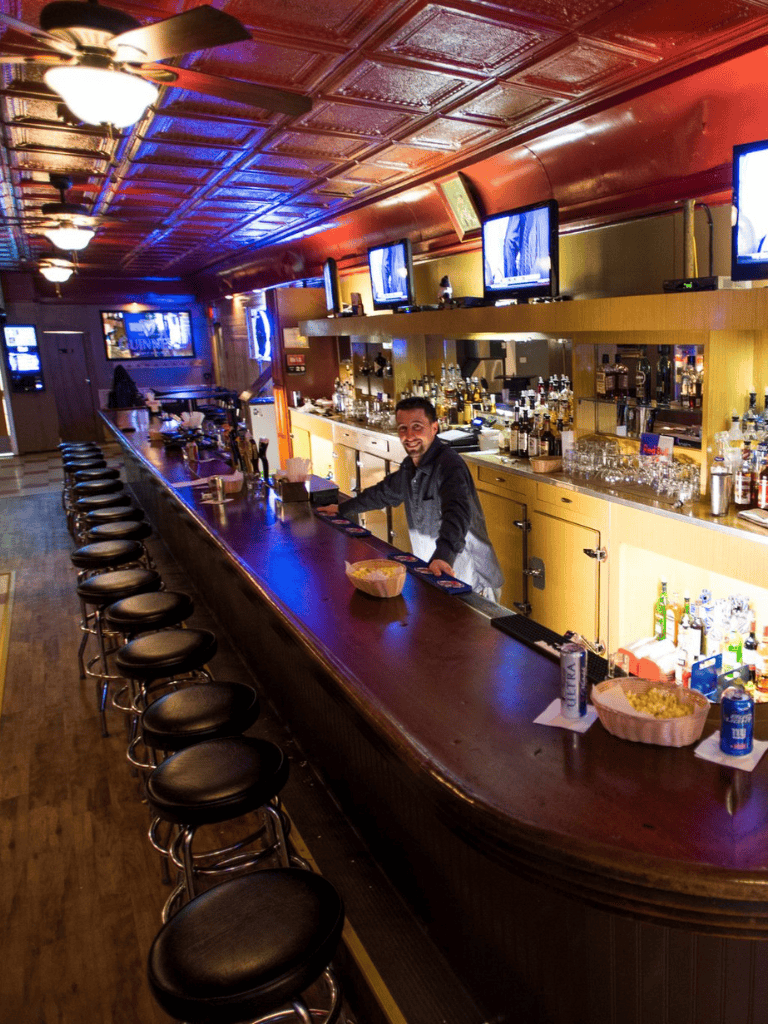 Cozy bar with multiple TV screens, well-stocked liquor shelves, and a friendly bartender for a great night out.