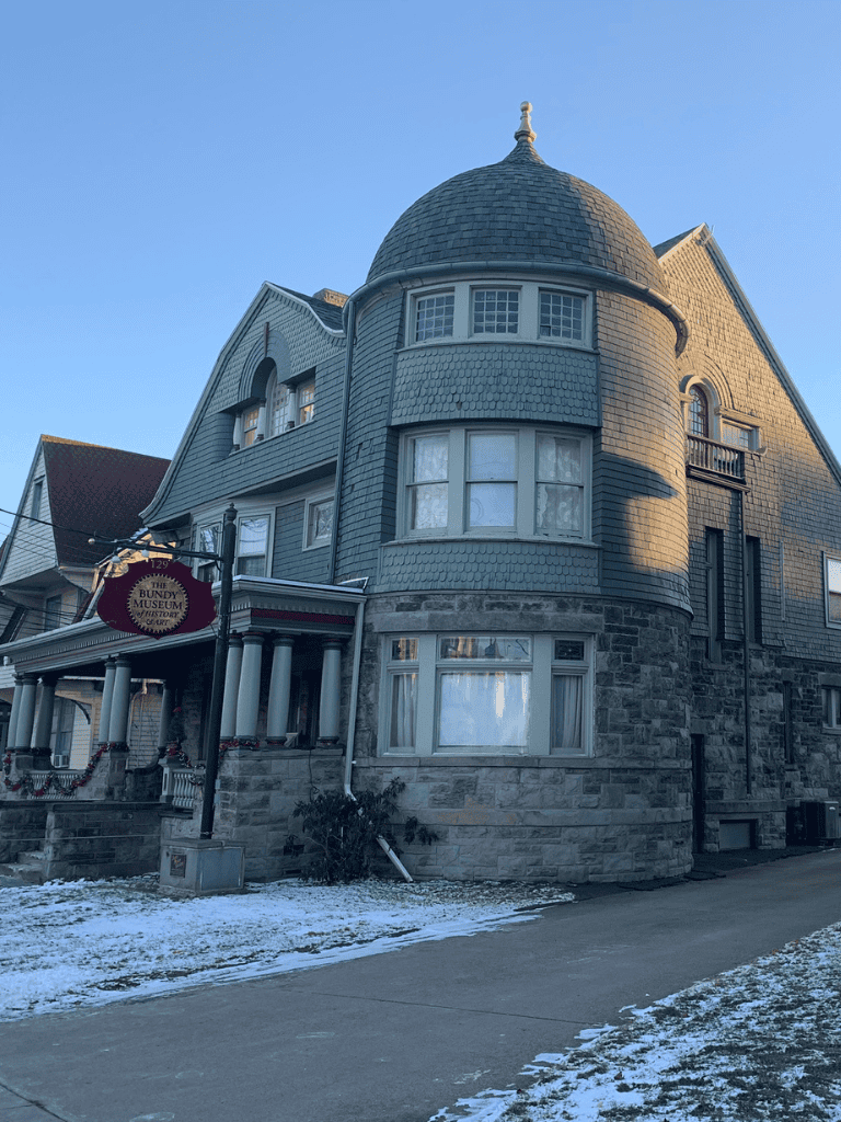Victorian-style house with rounded turret and historic museum sign in front, winter setting in the background.