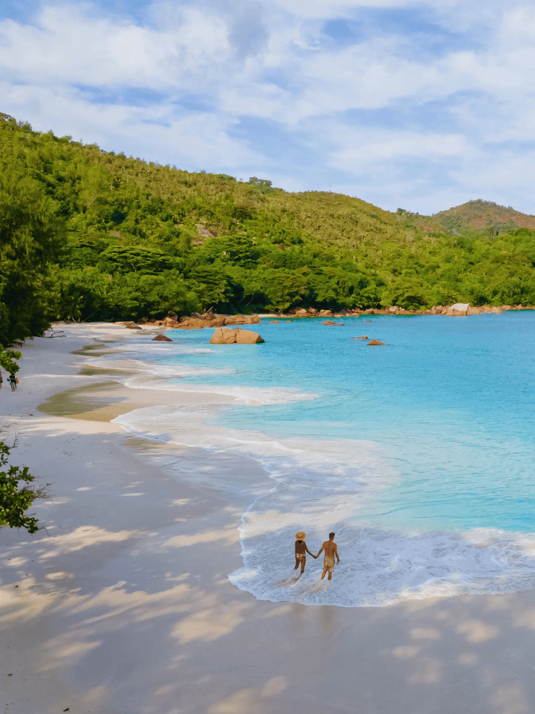 Tranquil beach with turquoise water, lush green hills, and two people holding hands in shallow waves.