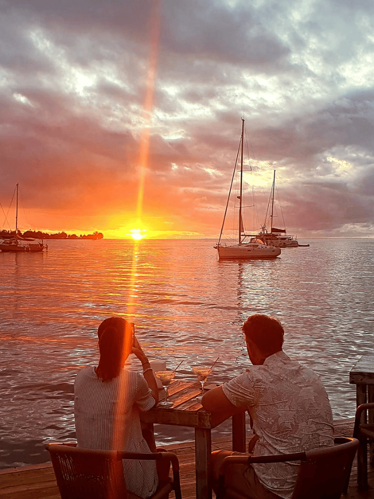 Sunset over the water with sailboats and a couple enjoying drinks by the dock, perfect for travel and leisure destinations.