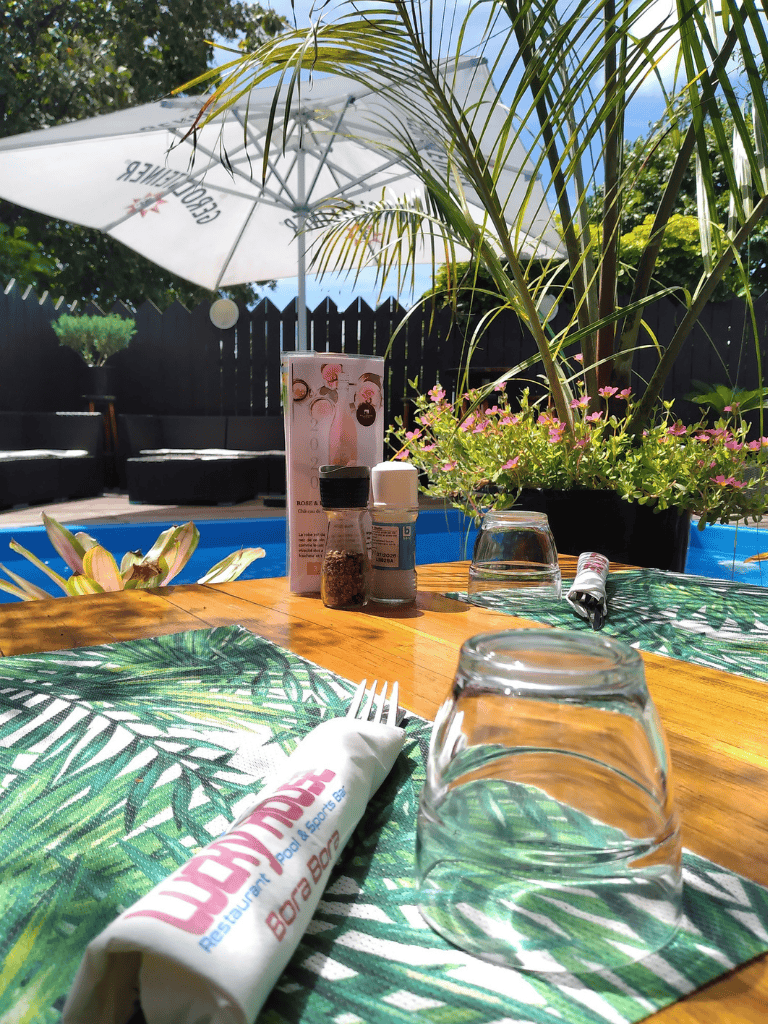 Bright outdoor dining setup with tropical plants, table settings, and an umbrella at QuestForDirections restaurant.
