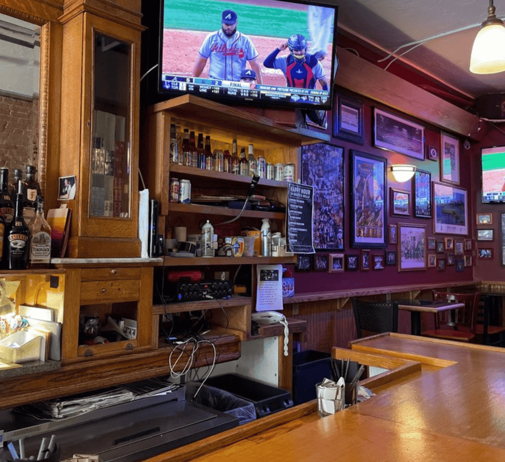 A cozy bar or restaurant interior with wooden decor, multiple framed pictures, and a large TV displaying a baseball game.