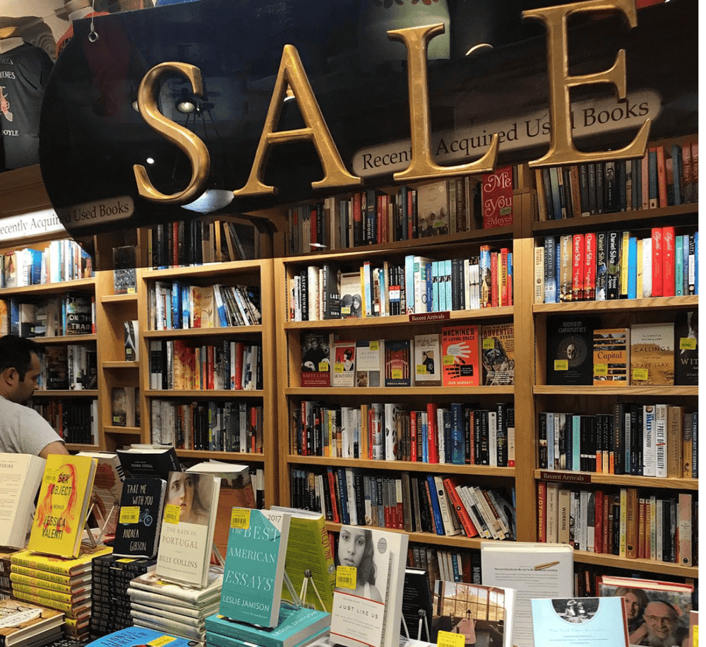 Used bookshelf at a bookstore with "SALE" sign, featuring a vast collection of used books and recent arrivals.