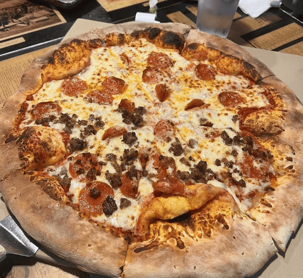Cheese pizza with pepperoni and sausage served on a wooden pizza peel at a restaurant.