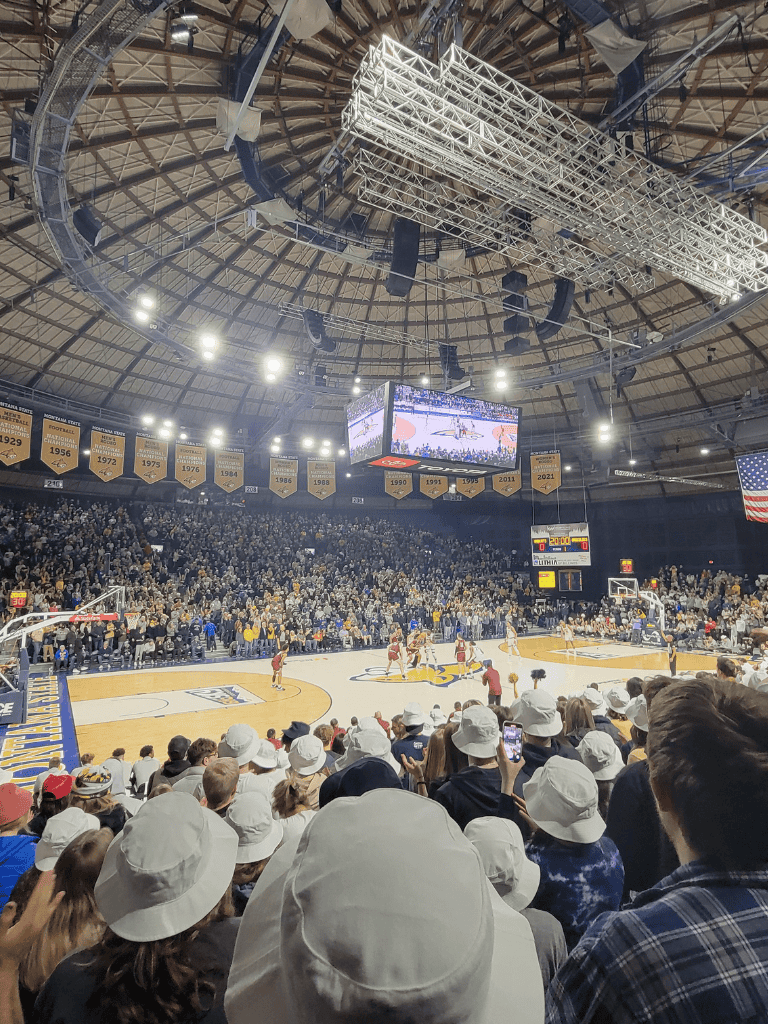Bright indoor sports arena during a basketball game with enthusiastic crowd and banners hanging from the ceiling.