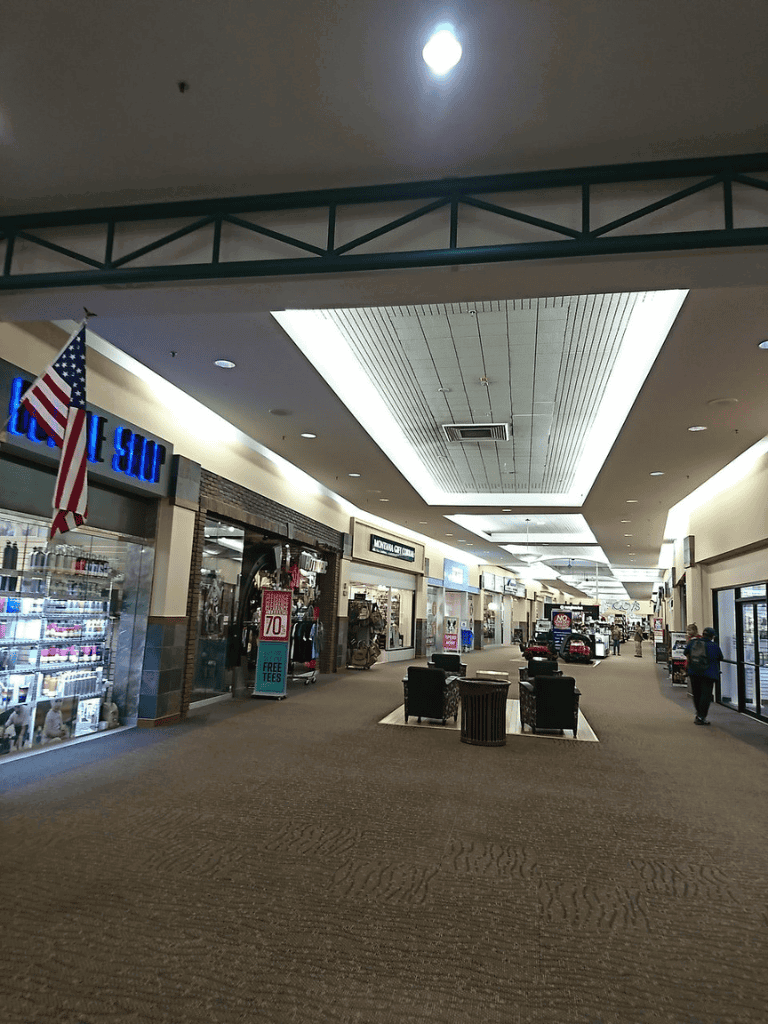 Wide view of shopping mall corridor with retail stores, seating area, and American flag for customer directions.