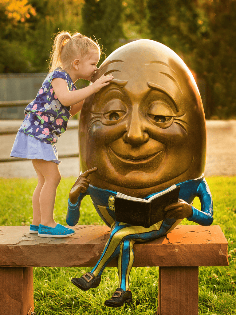 Playful young girl interacts with whimsical sun-themed sculpture reading a book in park outdoor scene.