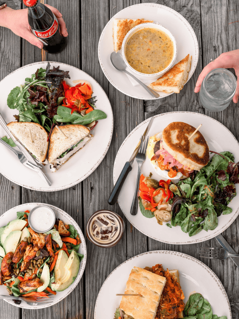 Freshly prepared sandwiches, salads, and beverages on a rustic wooden table, showcasing a diverse menu.