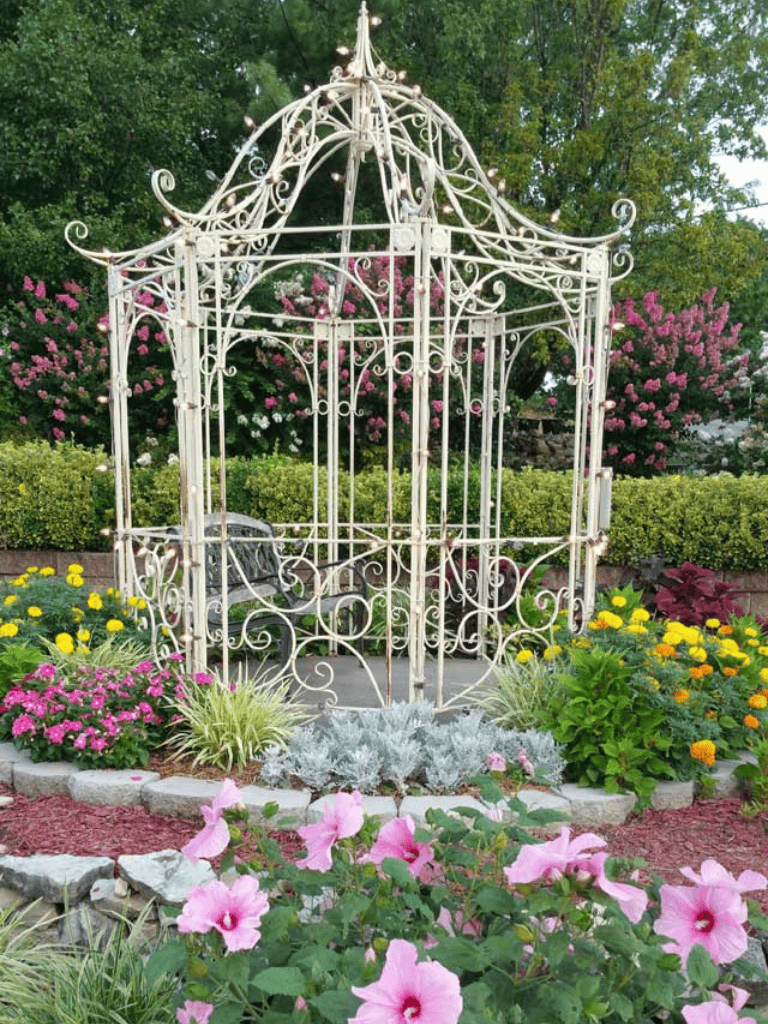Ornate white garden gazebo surrounded by colorful blooming flowers and lush green trees.