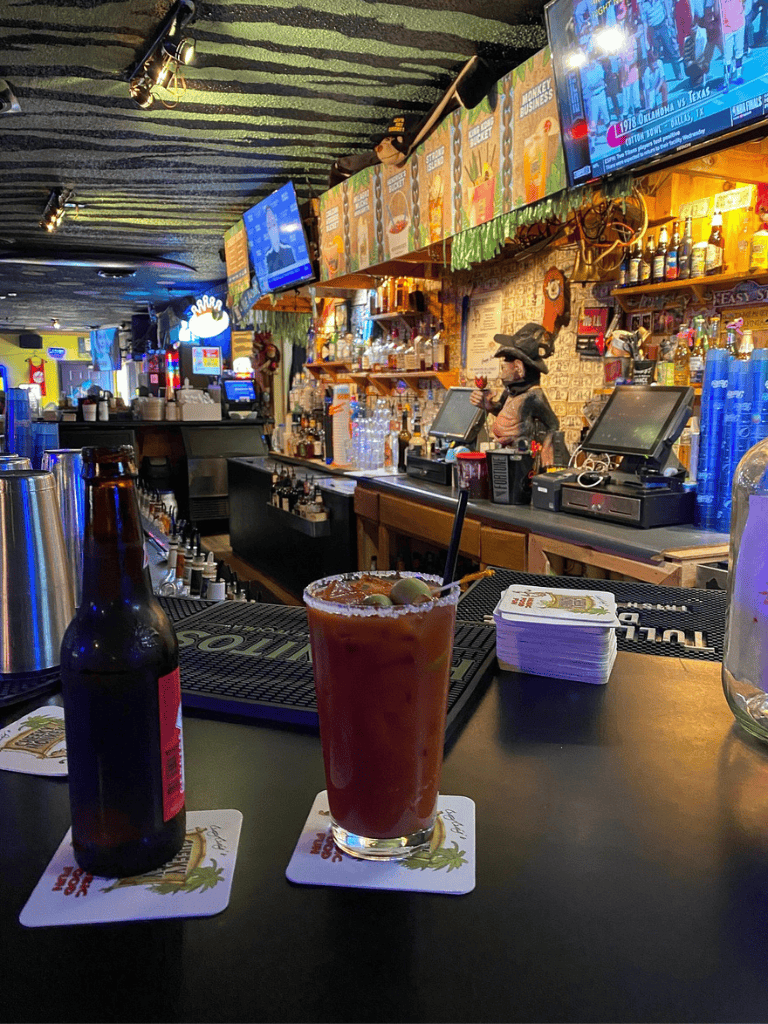 Refreshing bloody mary cocktail with a beer bottle on dark bar counter at colorful QuestForDirections bar, Texas.