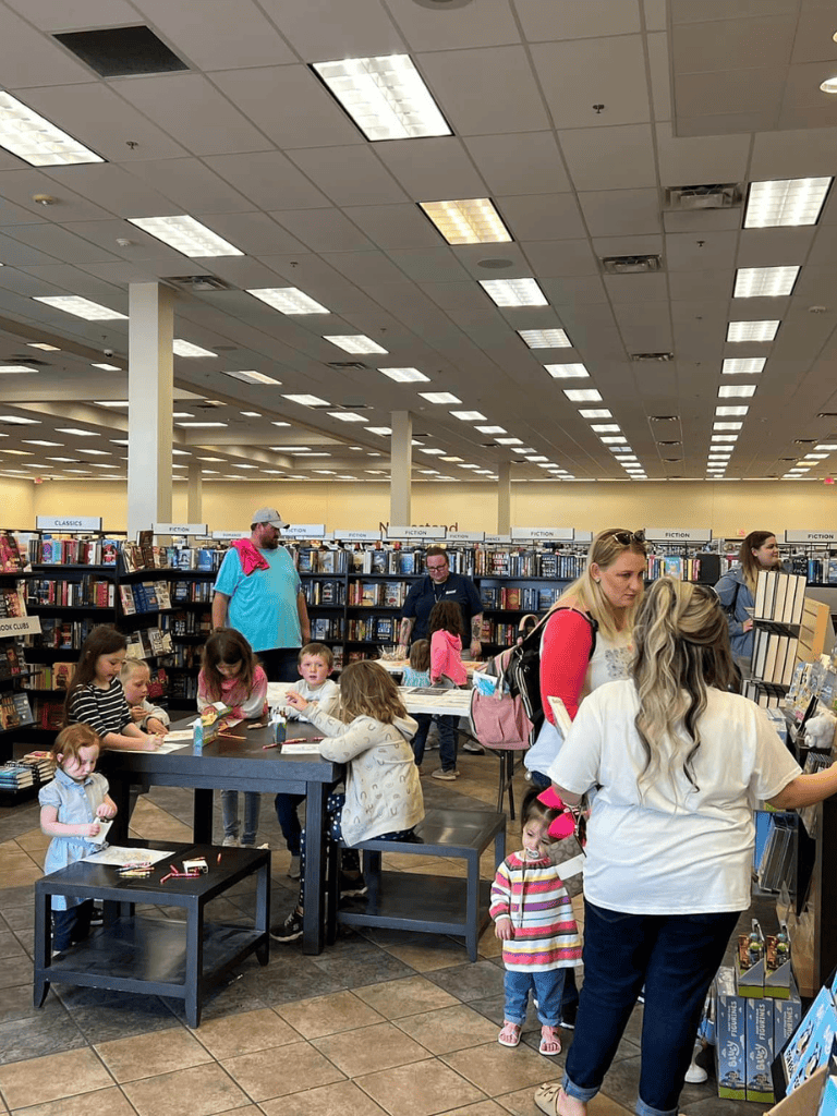 Bright indoor bookstore with children and adults browsing books and engaging in activities; well-lit with fluorescent ceiling lights.