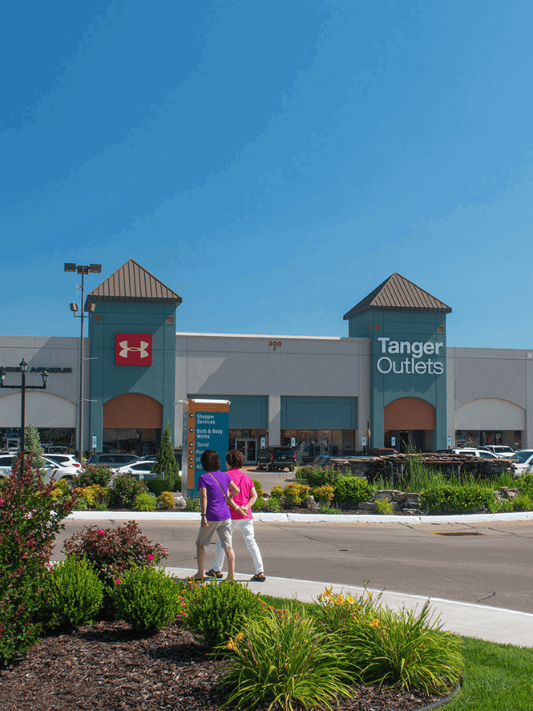 Bright outdoor scene of Tanger Outlets shopping center with two shoppers walking in the foreground.
