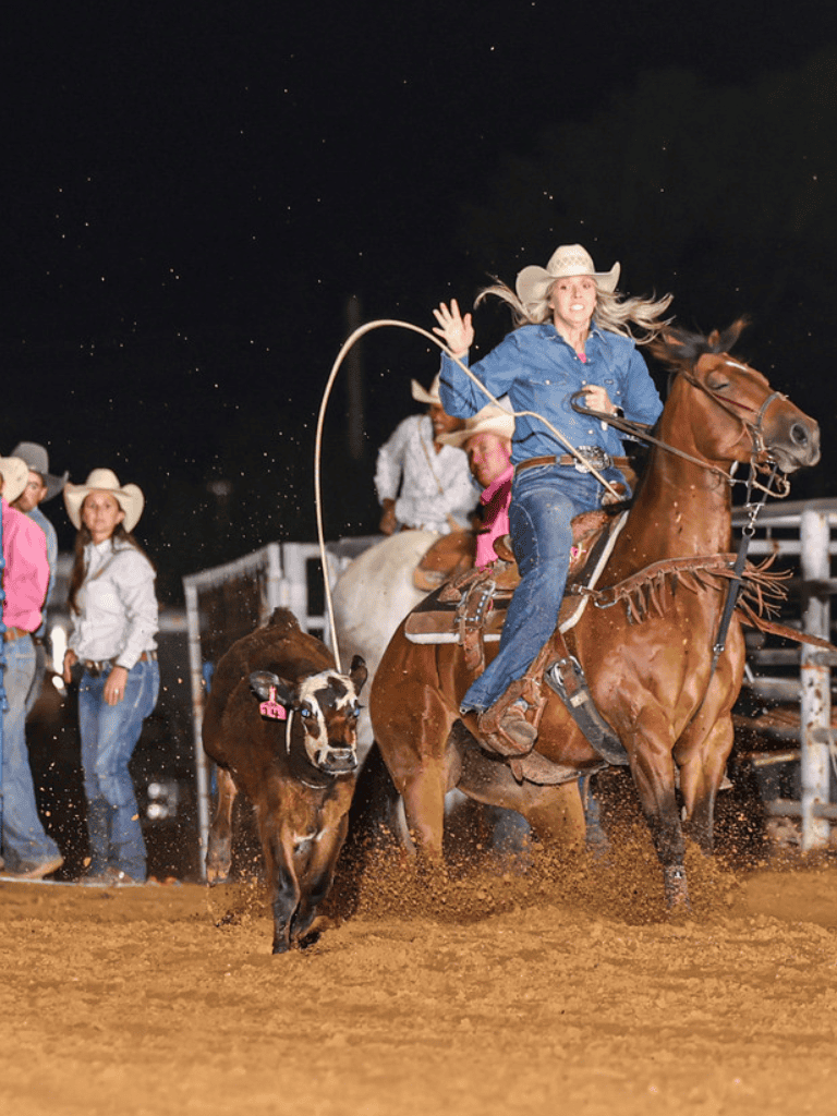 Rodeo cowgirl in blue shirt with lasso riding a bucking horse at night.
