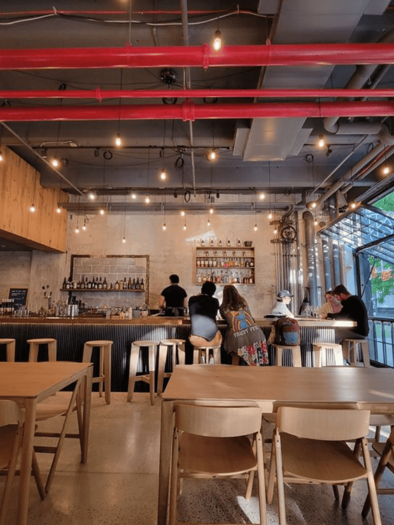 Industrial bar interior with high ceilings, exposed pipes, and modern lighting.