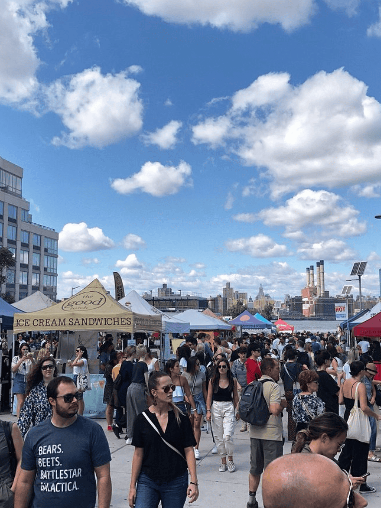 Busy outdoor street market with food stalls and crowds in New York City, sunny day with blue sky.
