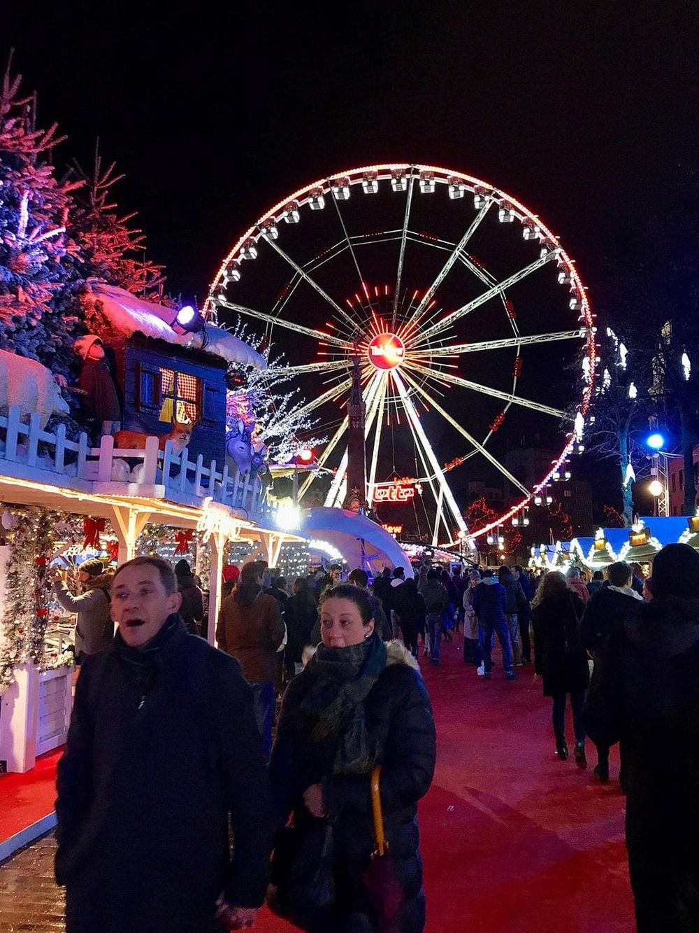 Bright Christmas market with a large illuminated Ferris wheel at night, festive atmosphere, holiday lights, and cheerful visitors.