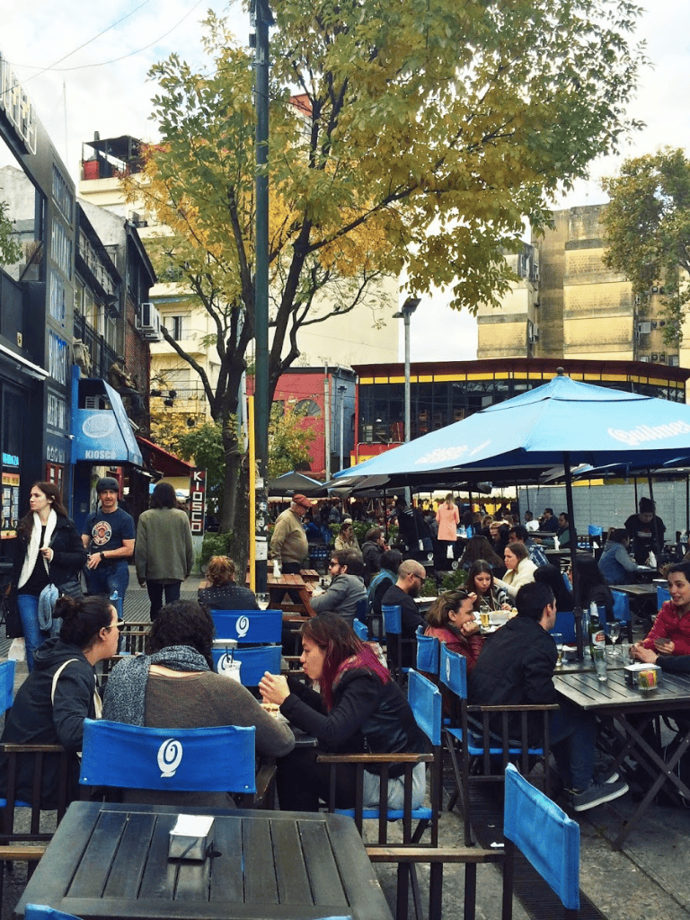 Outdoor dining area in a lively city street, crowded with people enjoying food and drinks.