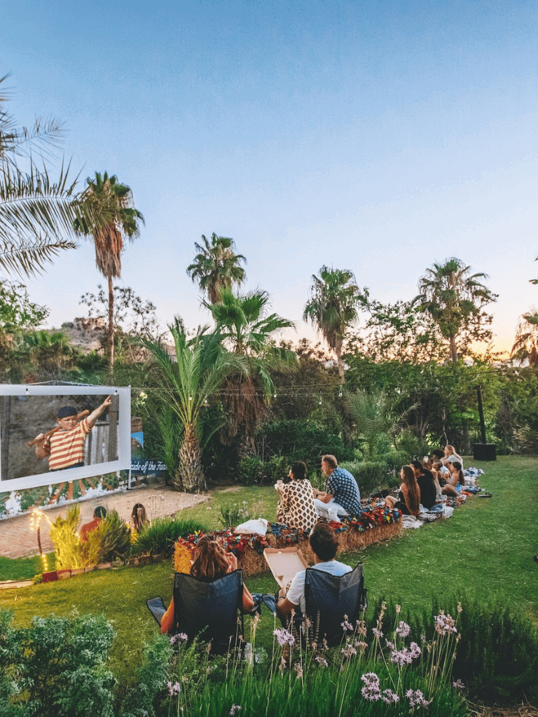 Outdoor movie night with people watching a film on a large screen in a lush garden setting.