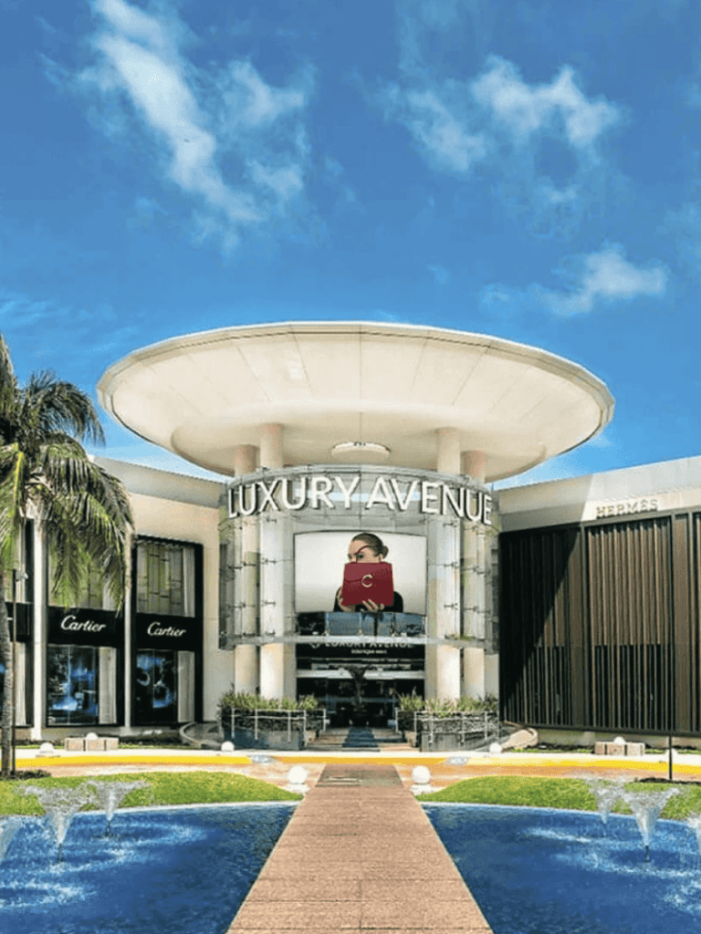 Luxury shopping mall exterior with high-end stores like Cartier and Hermès, modern architecture, and water fountains under a blue sky.