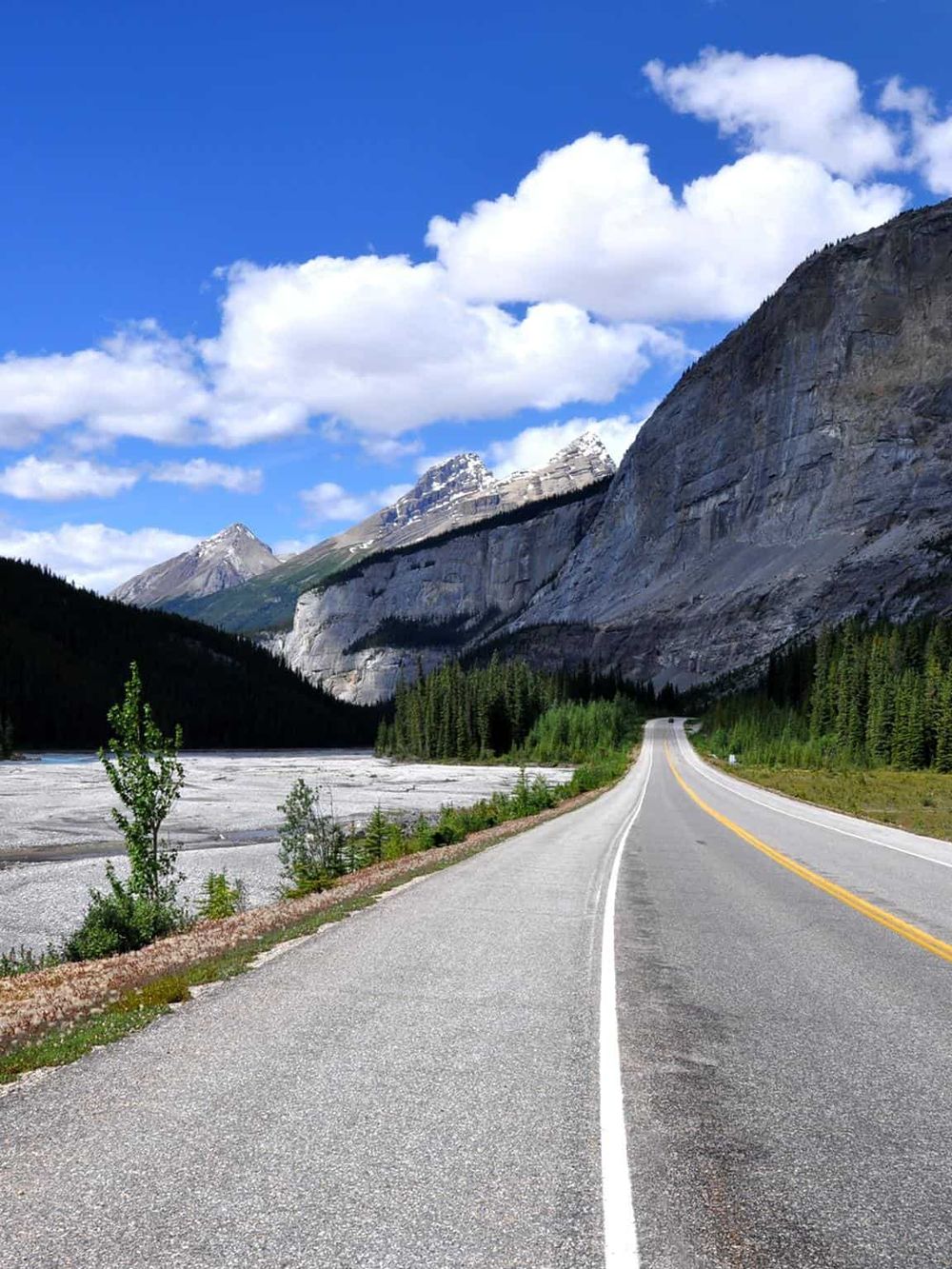 Serene mountain road surrounded by lush forests and striking peaks under a bright blue sky.