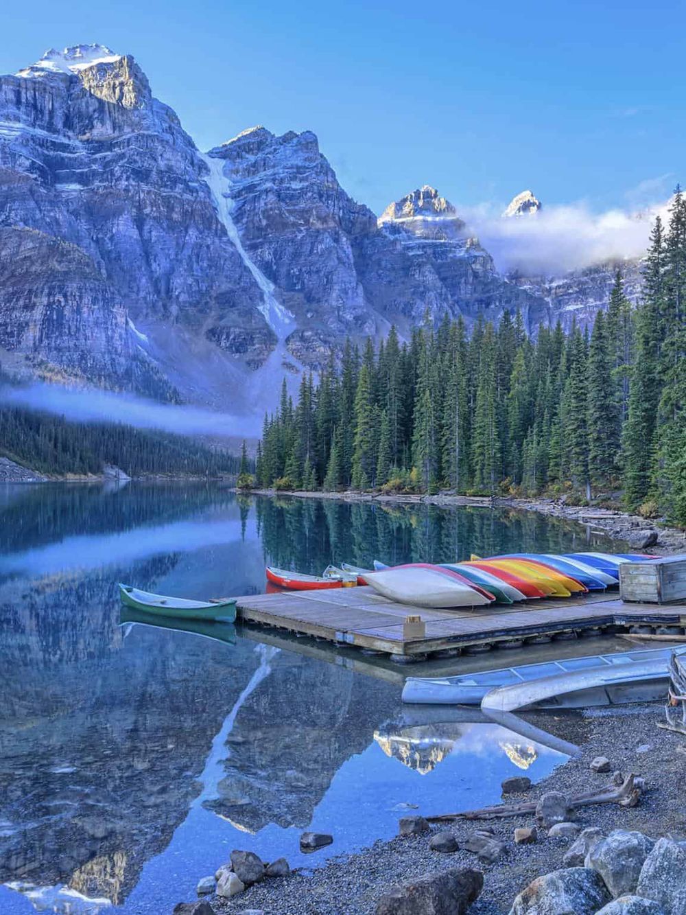 Serene mountain lake with colorful kayaks, surrounded by pine trees and towering snow-capped peaks.