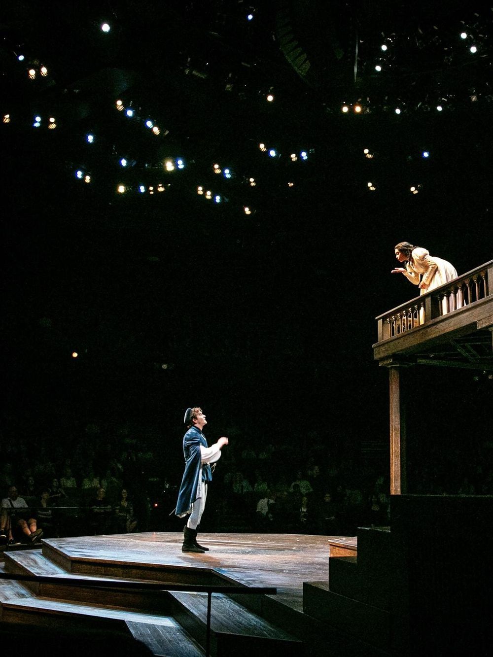 Theater performance scene with actors on stage, dramatic lighting, and audience in background.
