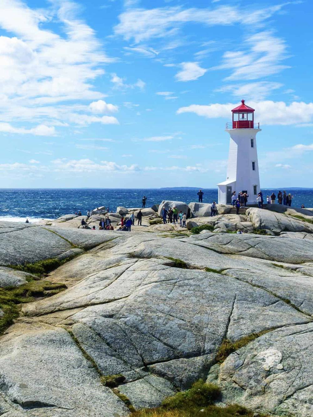 Lighthouse on rocky shoreline with visitors, ocean view and blue sky, popular sightseeing spot.