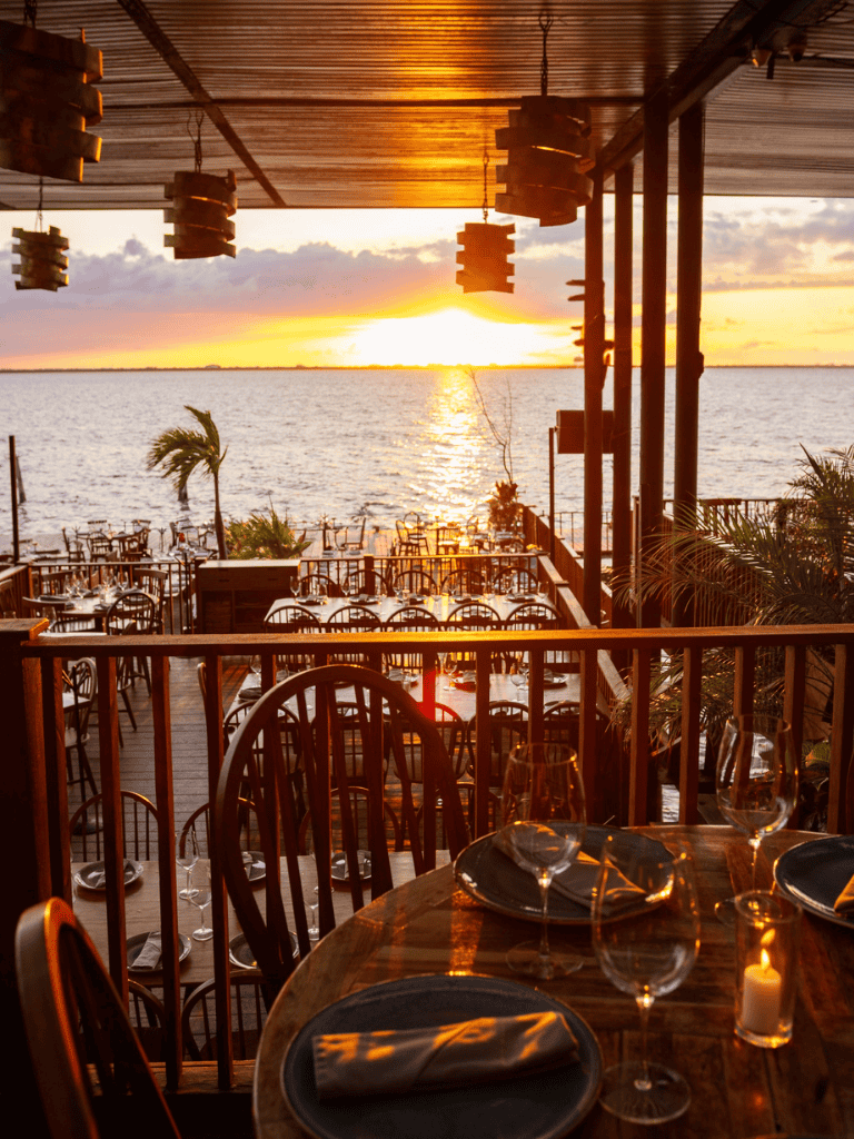 Sunset over ocean at beachfront restaurant with outdoor dining tables and tropical decor.