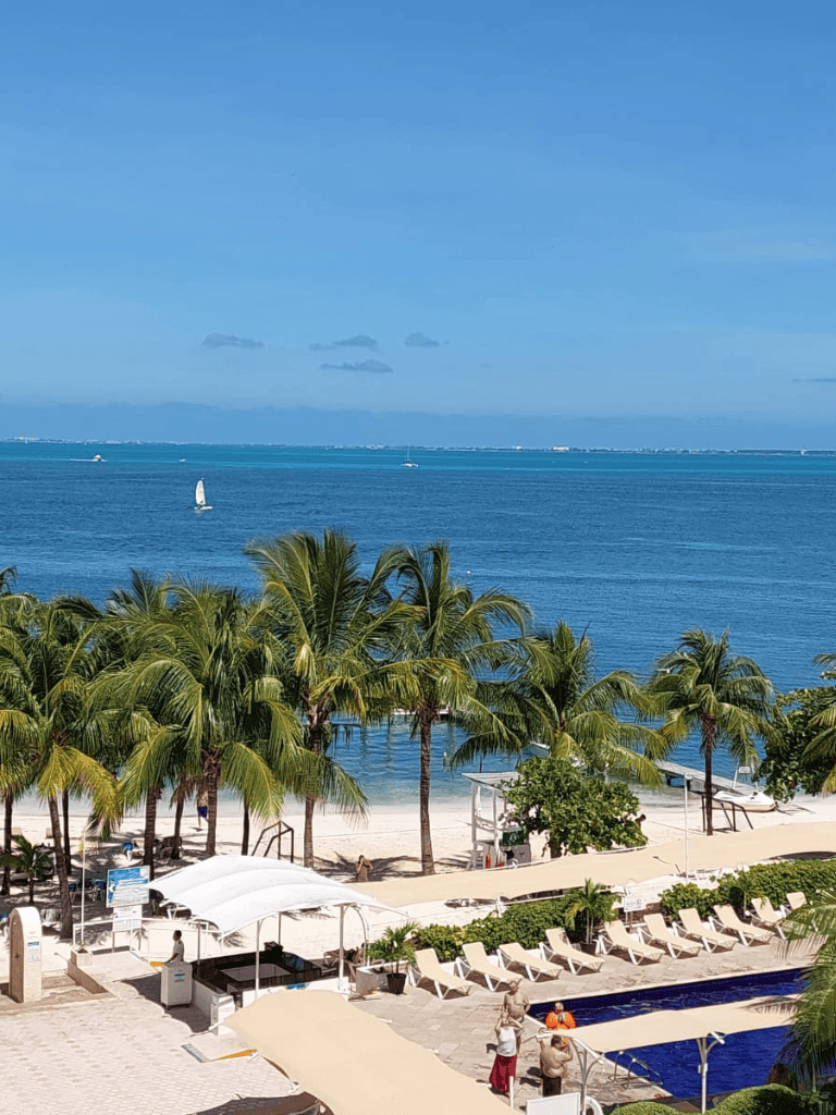 Relaxing beach view with palm trees, poolside lounge chairs, and sailing boats on calm ocean waters.