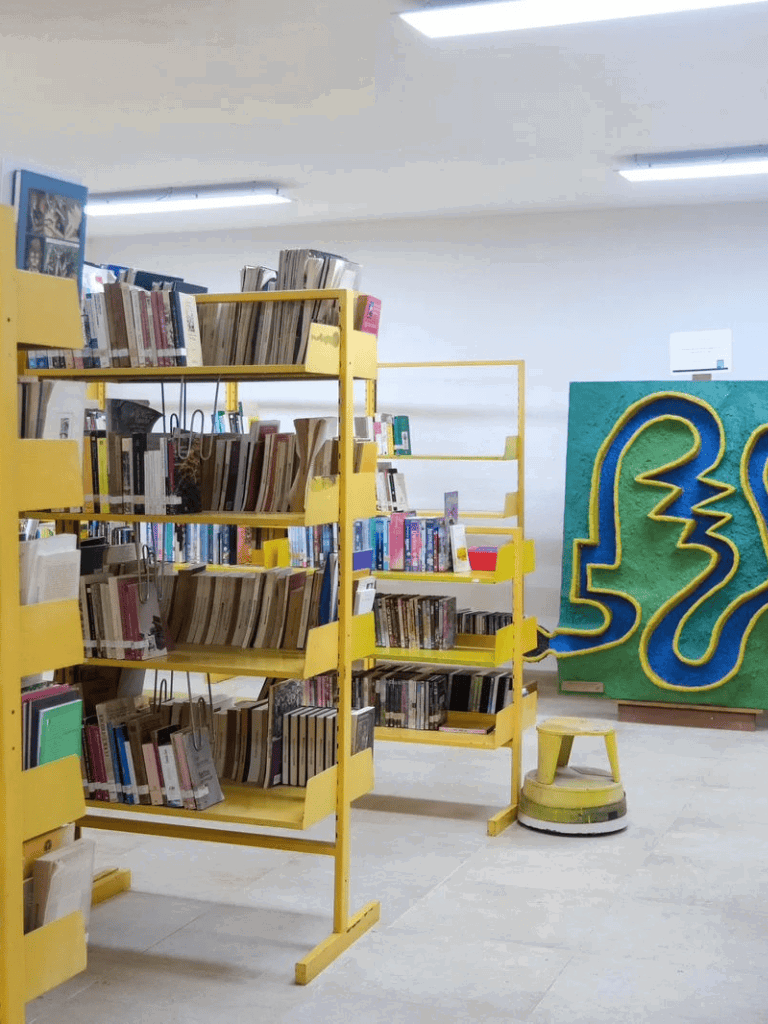 Bookshelves with diverse books in a library setting, featuring bright yellow racks and artwork in the background.