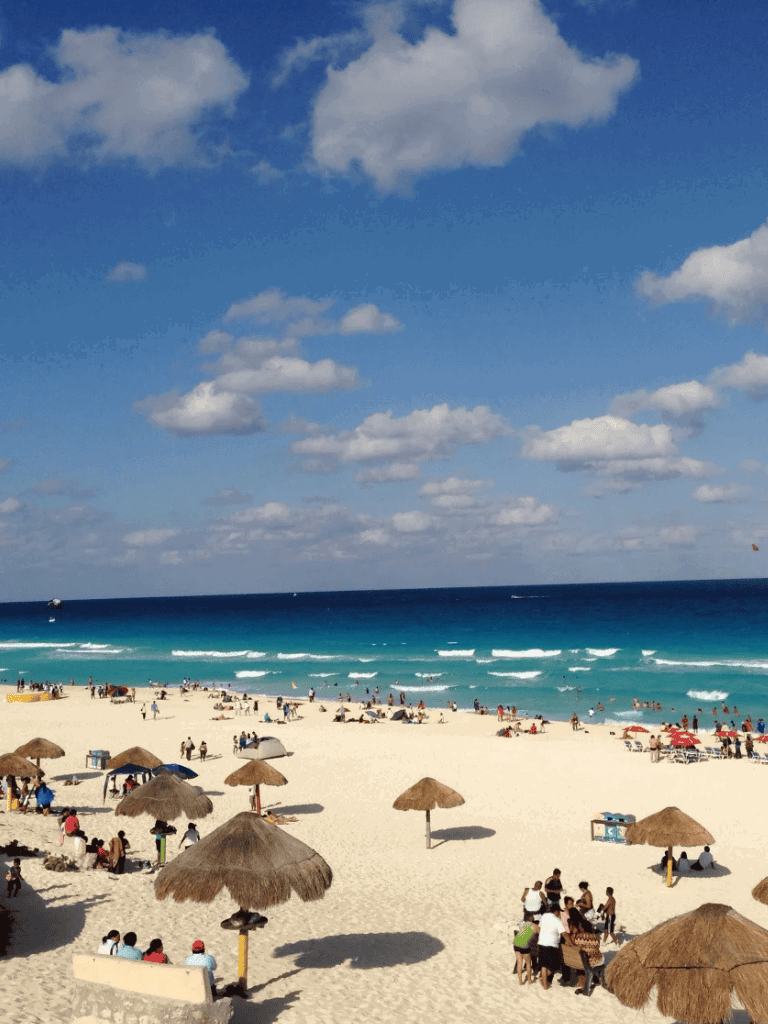 Vibrant beach scene with umbrellas, surfers, and blue ocean under a partly cloudy sky.