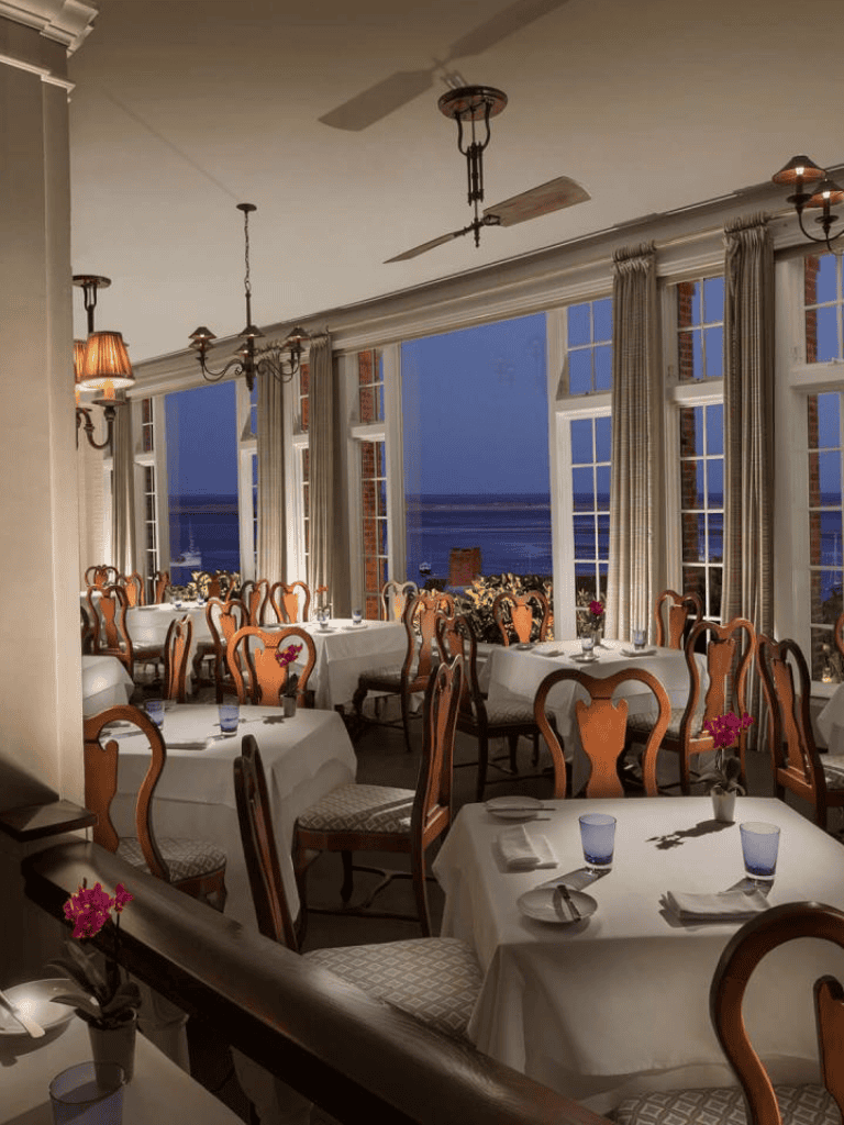 Cozy waterfront restaurant interior with elegant wooden chairs, white tablecloths, and ocean views at sunset.