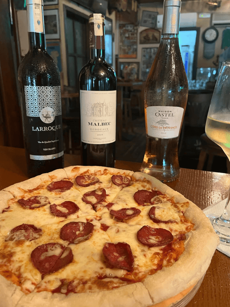 Aged wine bottles, a pepperoni pizza, and a glass of white wine in a cozy café setting.
