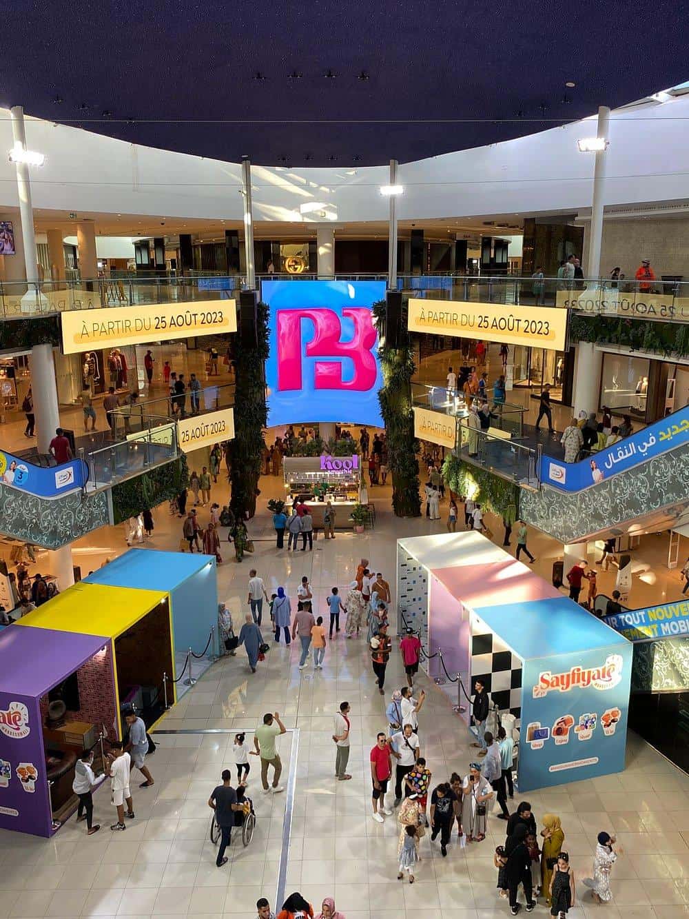 Bright shopping mall interior with digital signage, shoppers, and multiple levels, promoting an event starting August 25, 2023.