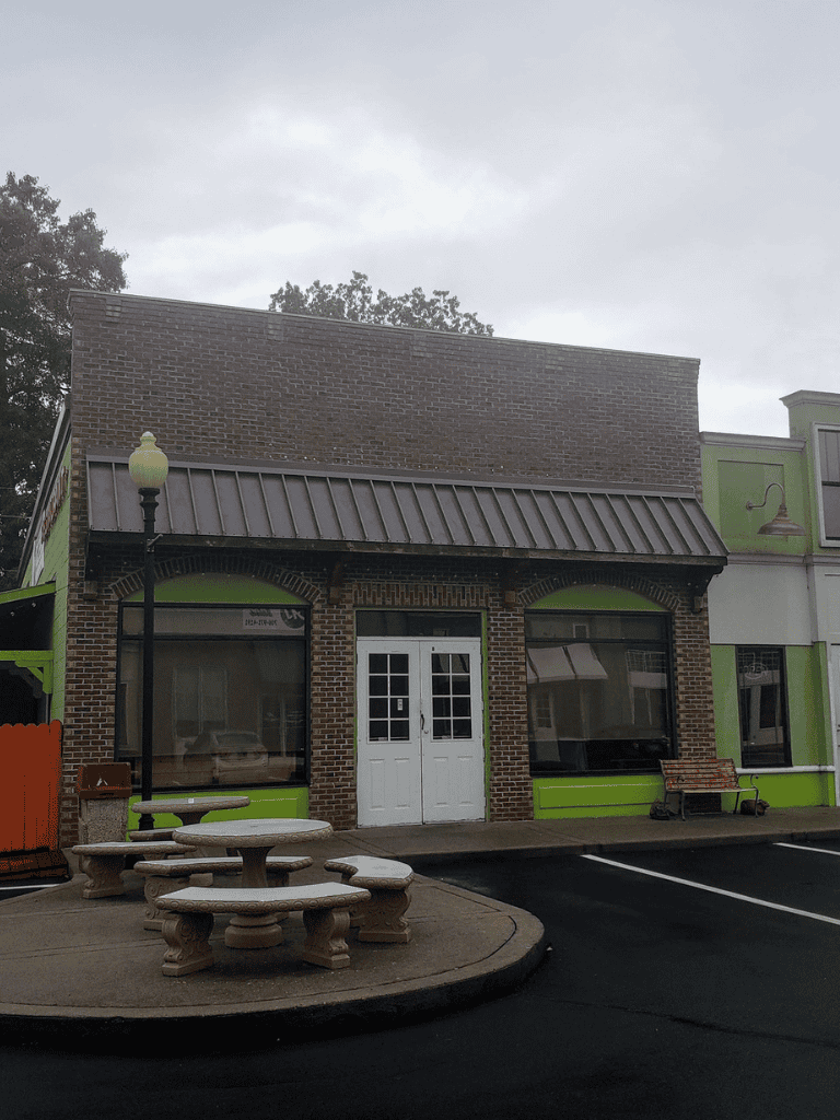 Small brick building with green accents, outdoor seating, and a parking lot, ideal for local businesses or restaurants.