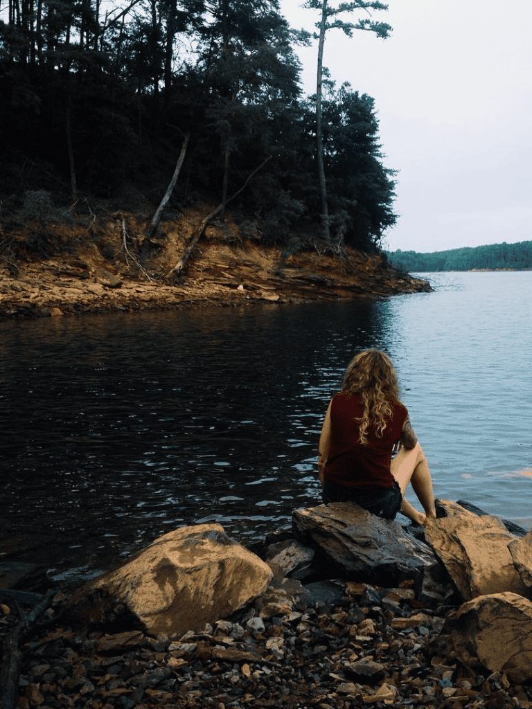 Secluded lakeside scene with woman sitting on rocks, surrounded by nature and trees.