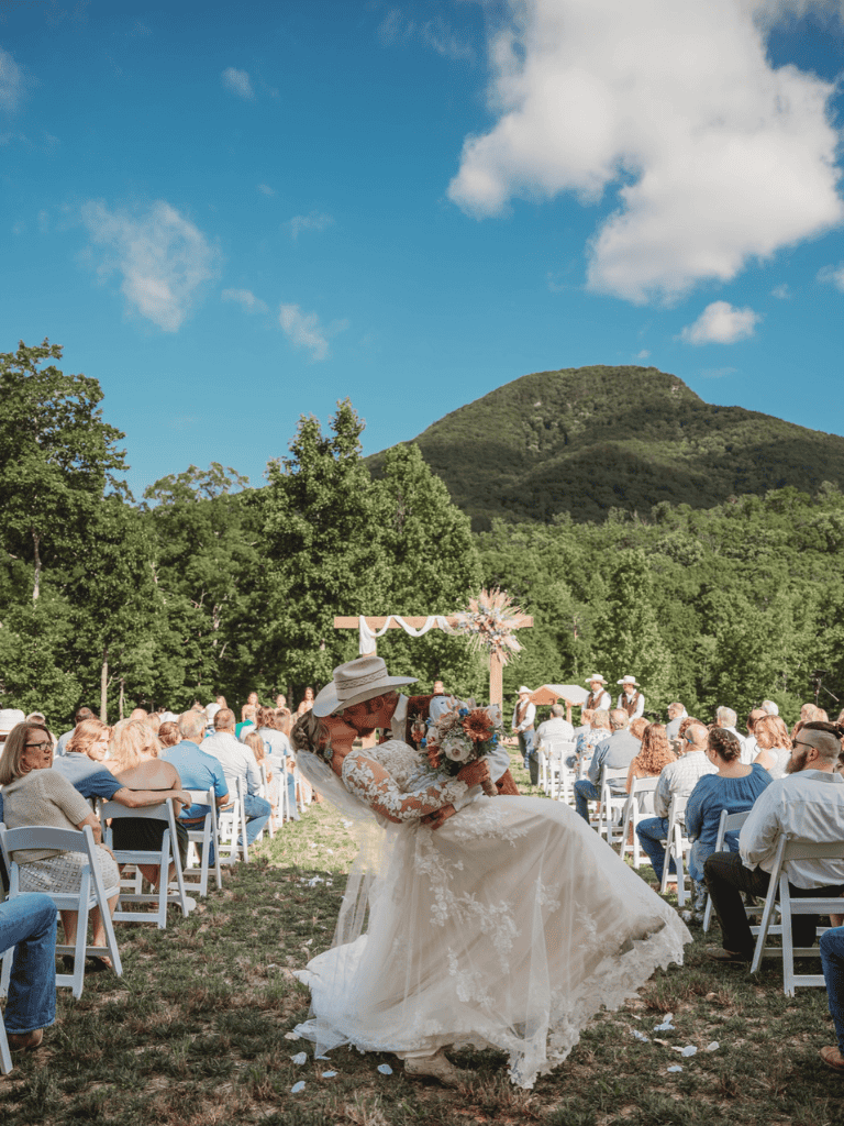 Happy couple at outdoor wedding ceremony, joyful bride and guests, scenic mountain backdrop, sunny day.