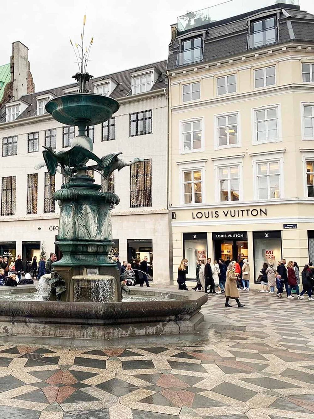 Intricate city fountain with pedestrians outside Louis Vuitton store in a busy shopping district.