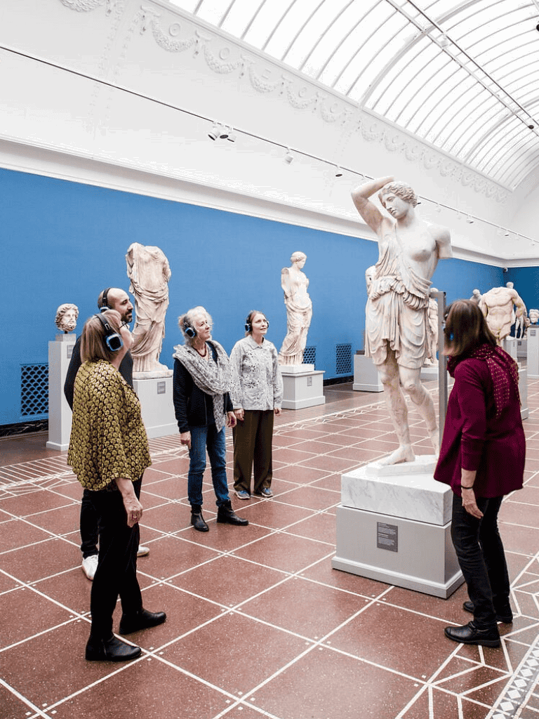 Ancient Greek sculptures exhibit at museum with visitors listening to audio guides.