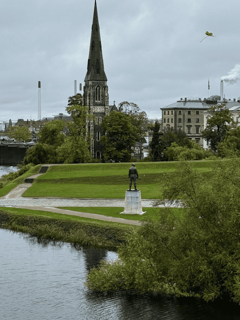 Historic church and statue in a lush park along a river, city skyline with industrial chimneys in the background.