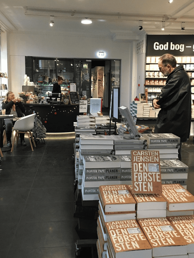 Aisle of books at a bookstore with customers browsing and a cafe counter in the background.