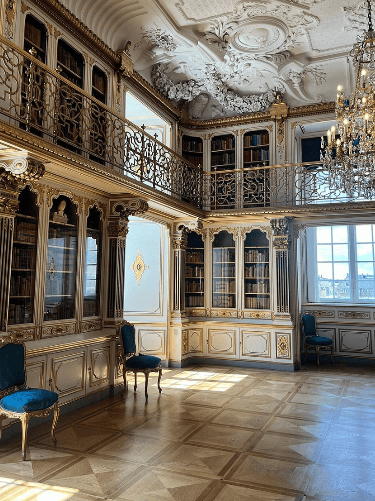 Opulent classical library with gold accents, chandeliers, and extensive book collection.