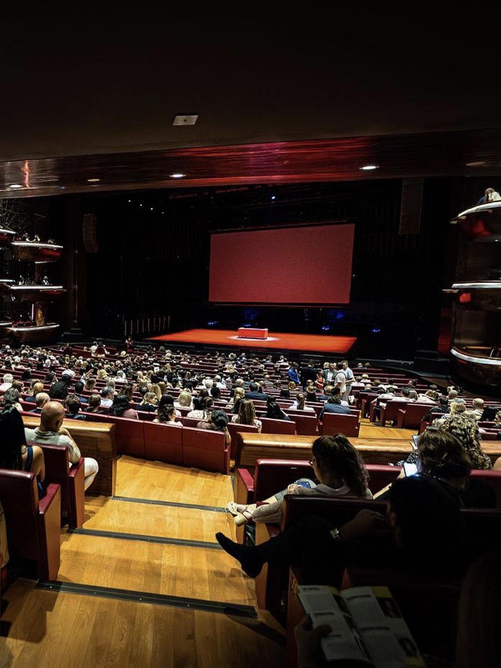 Aerial view of a crowded auditorium during a conference or event, with a large screen and stage at the front.