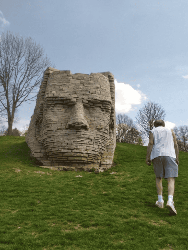 Ancient stone head sculpture on grassy hill with person walking nearby, scenic outdoor art installation.