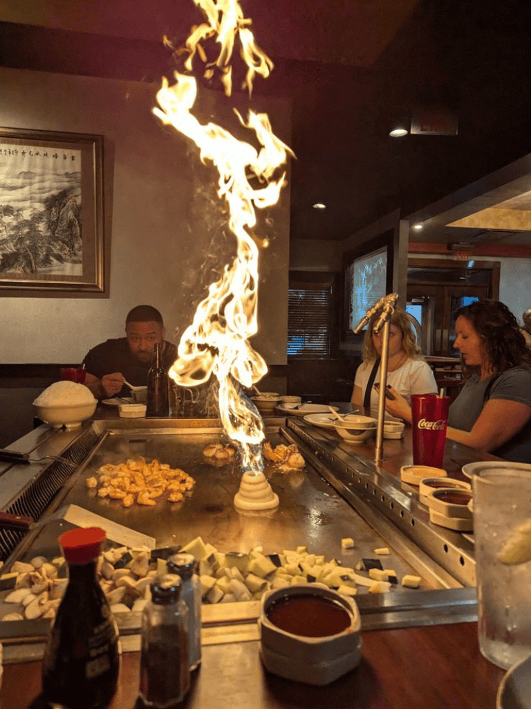 Vibrant teppanyaki restaurant with flames and lively dining atmosphere, showcasing delicious sushi and Asian cuisine.