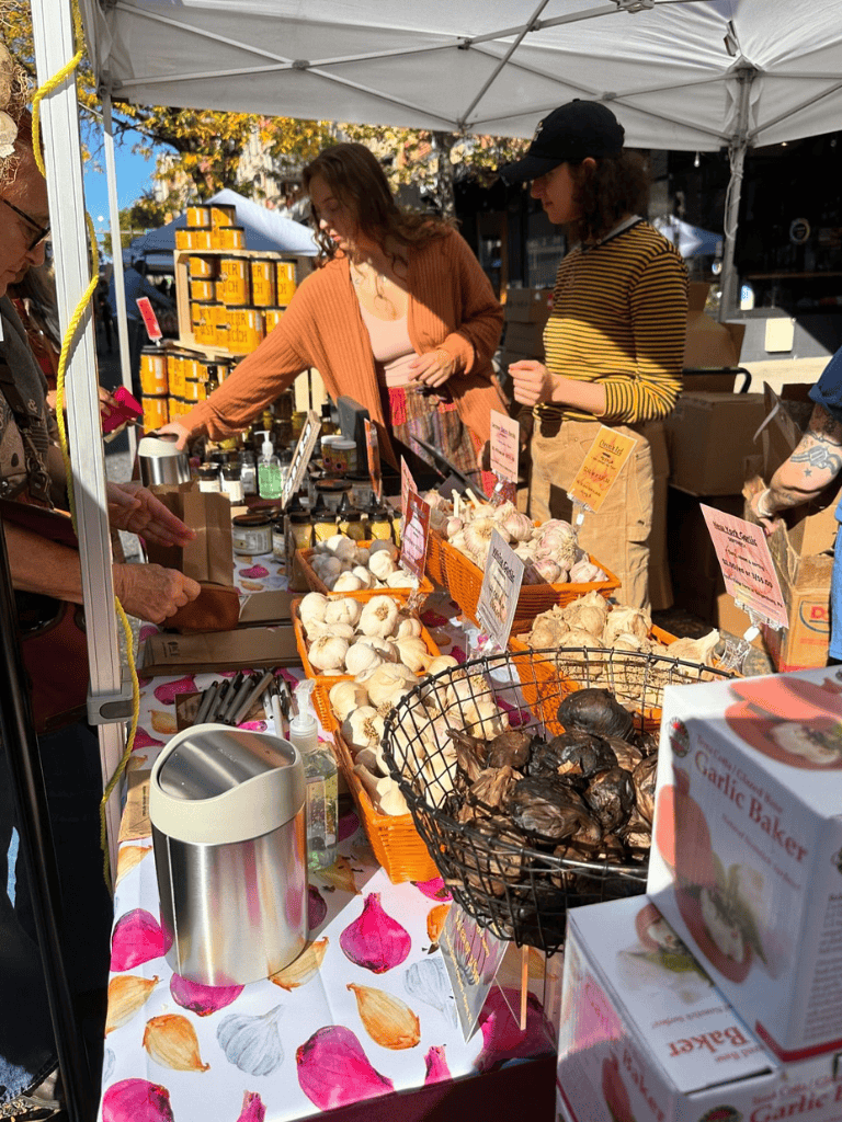 Fresh garlic and produce stall at outdoor market, sunny day, shoppers browsing local vendors, colorful displays, community event.