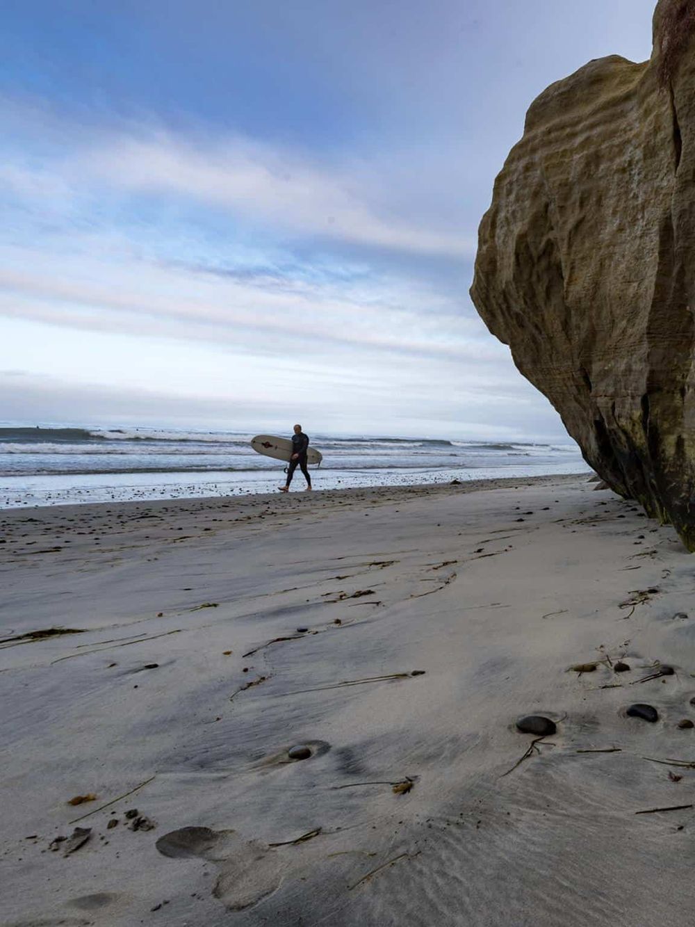 Surfer walking on the beach with surfboard and large rock formation, coastal scenery.