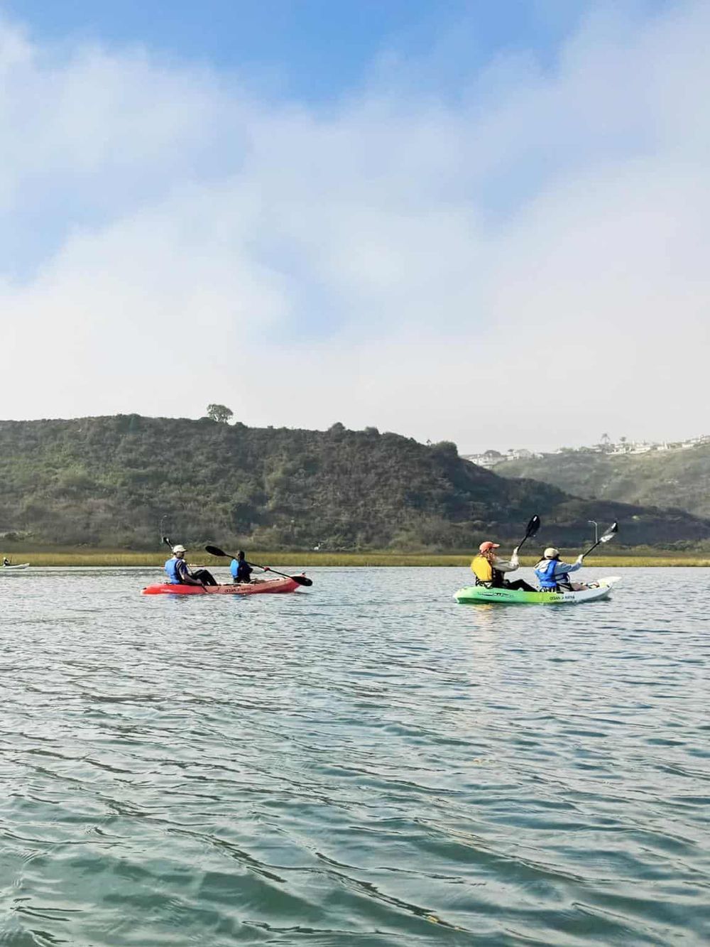 Kayakers paddling on calm water with scenic hills and blue sky in the background.