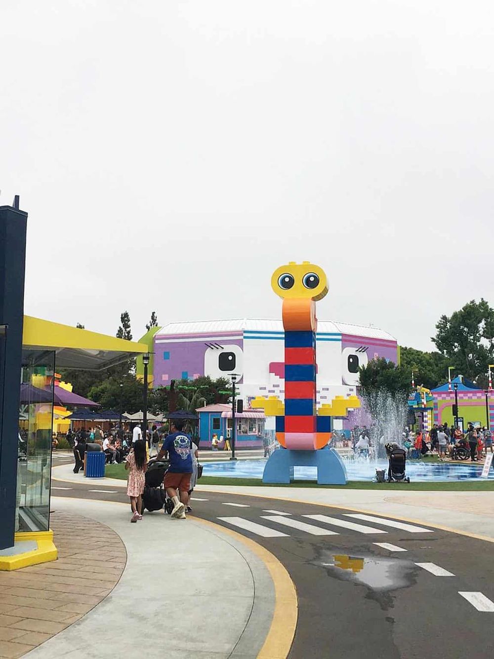 Colorful Lego-themed amusement park with giant Lego character fountain and family enjoying the attractions.